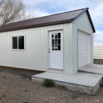 A small white metal shed with a brown roof stands quietly on a concrete slab, featuring a door with a window and an adjacent garage door. Sparse grass grows around the structure, while in the background, a leafless tree adds an elegant touch to this picturesque scene—perfect for Temp Blog Post 1.