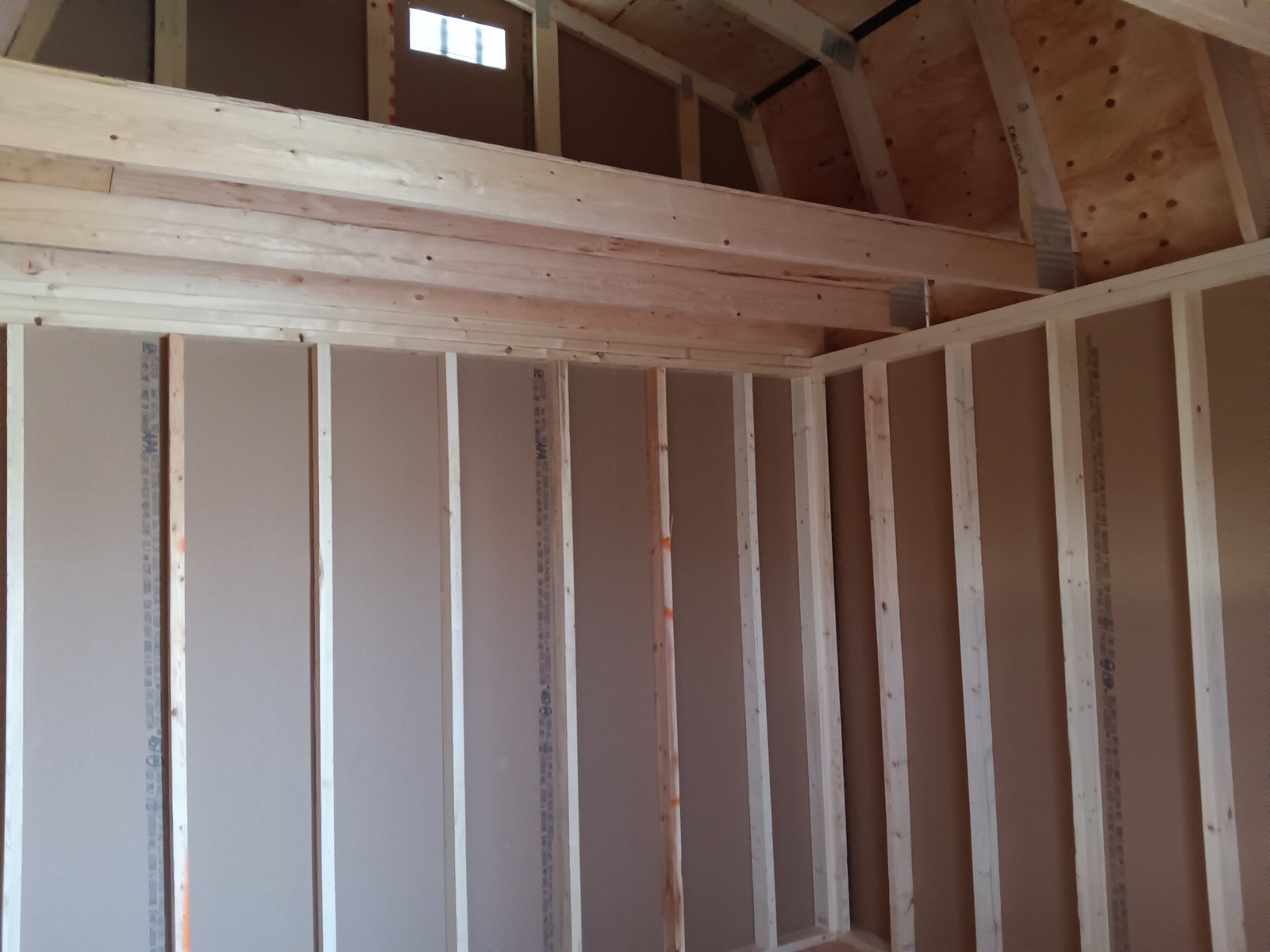 Interior view of the Lofted Dutch Barn - 2114 under construction. The image features exposed wooden beams and studs, along with partially installed drywall. A small window near the top allows natural light to illuminate this rustic space, showcasing the craftsmanship involved in creating this barn.