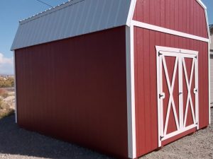 A Lofted Dutch Barn - 2113 shed featuring a white roof and trim rests on a gravel surface with its doors sealed, all beneath a clear blue sky. Power lines trace the horizon in the background, enhancing the rural charm.