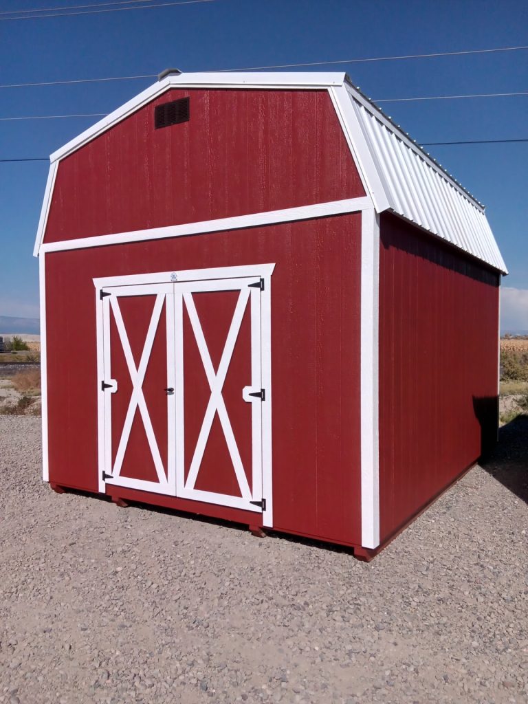 A Lofted Dutch Barn - 2113, featuring white doors and trim, rests on a gravel surface beneath the clear blue sky. Its charming peaked roof stands out as power lines trace the background.
