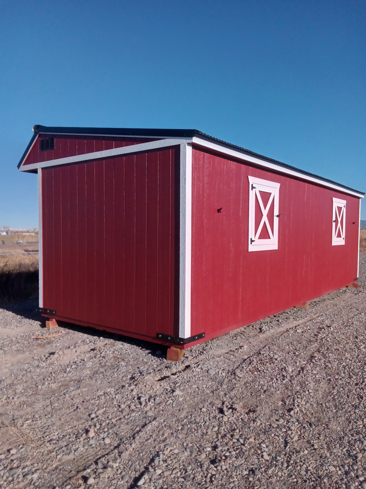 A red, barn-style shed with white trim and cross-braced windows sits on a gravel surface under a clear blue sky, reminiscent of the idyllic scene captured by the product Horseman - 2303.