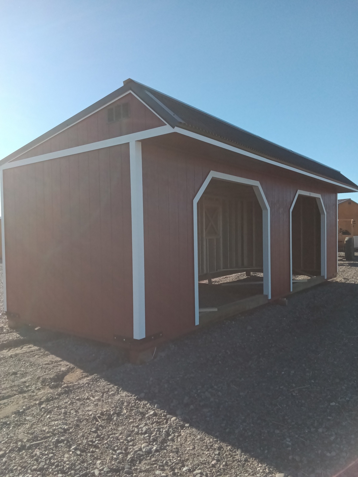 The Horseman - 2303, a red wooden shed with white trim and an open interior that evokes the charm of a rustic horseman's retreat, is nestled on a gravel lot beneath the vast blue sky.