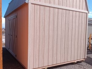 The Lofted Dutch Barn - 2368, featuring a crossbeam door and metal roof, is a charming structure in beige wood. It stands elegantly on a gravel surface beneath the clear blue sky.