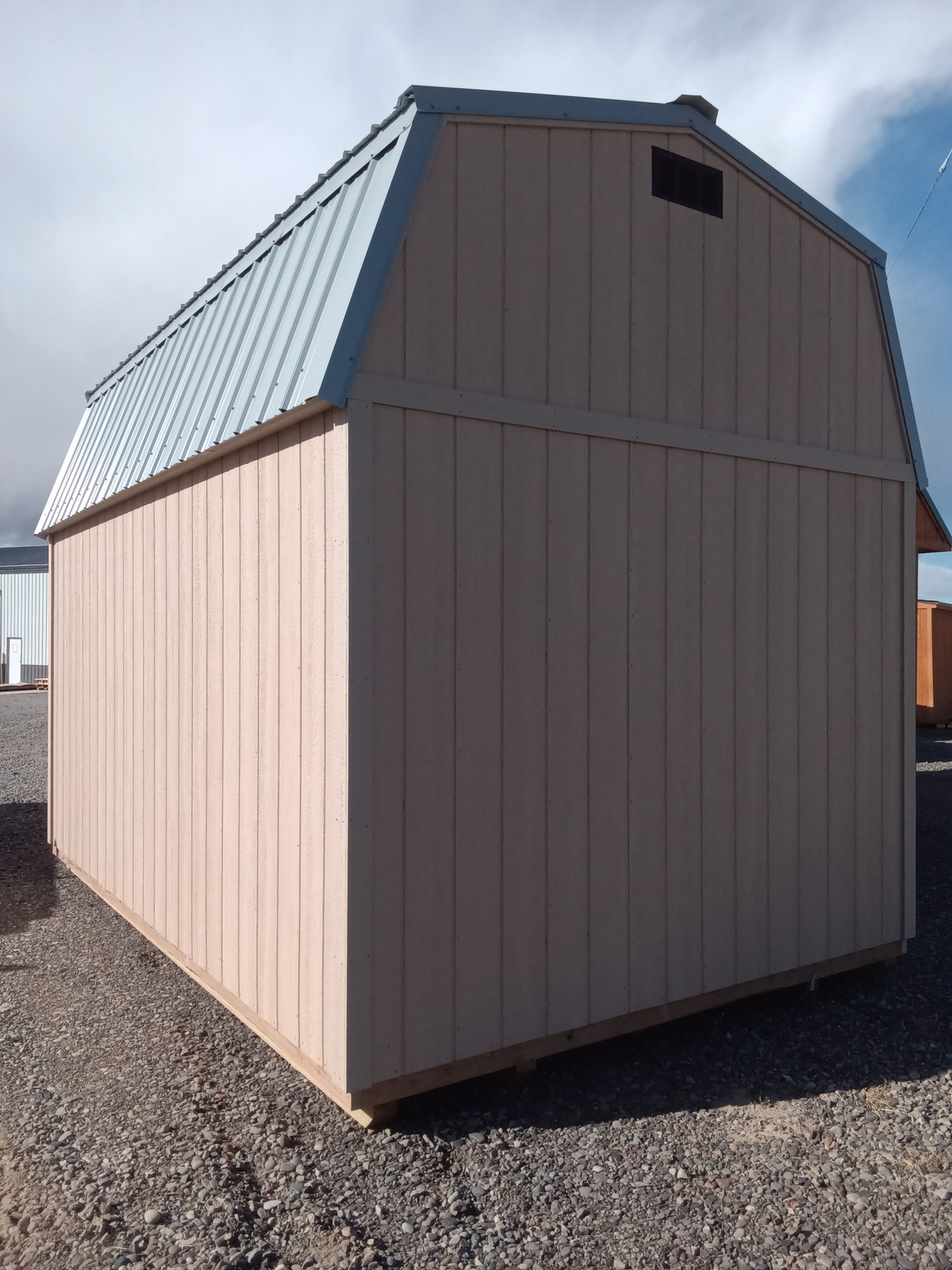 A Lofted Dutch Barn - 2368, small and light-colored, with a wooden structure and metal roof, stands on a gravel surface under a partly cloudy sky. It features the distinctive lofted design and includes a vent near the peak of the roof. Similar barns can be seen in the background, enhancing this picturesque rural setting.