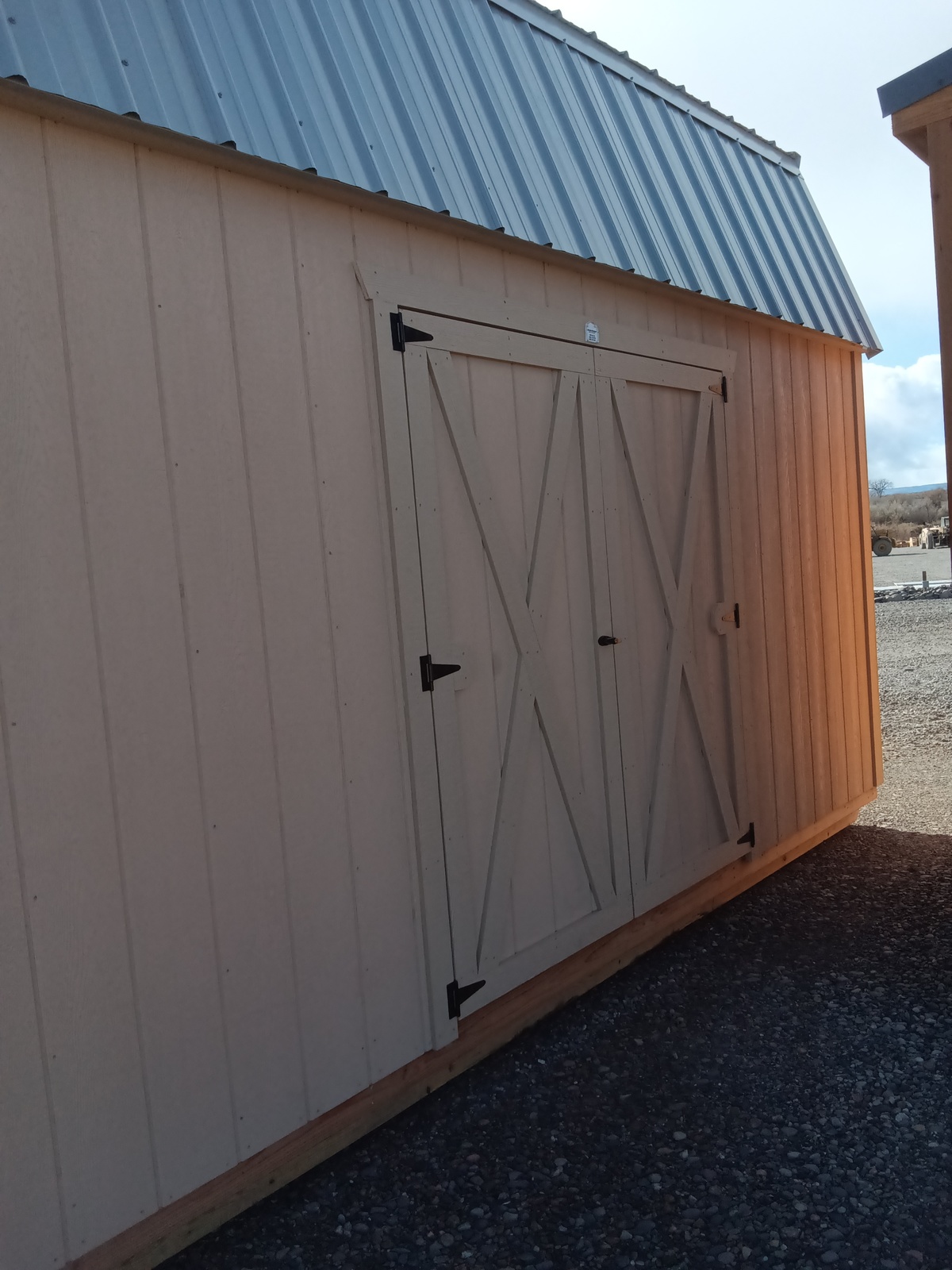 The Lofted Dutch Barn - 2368, featuring a tan wooden exterior, metal roof, and X-braced double doors, exudes a charming rustic appeal. Positioned on a gravel base beneath an open sky, it stands alongside another partially visible structure to the right.