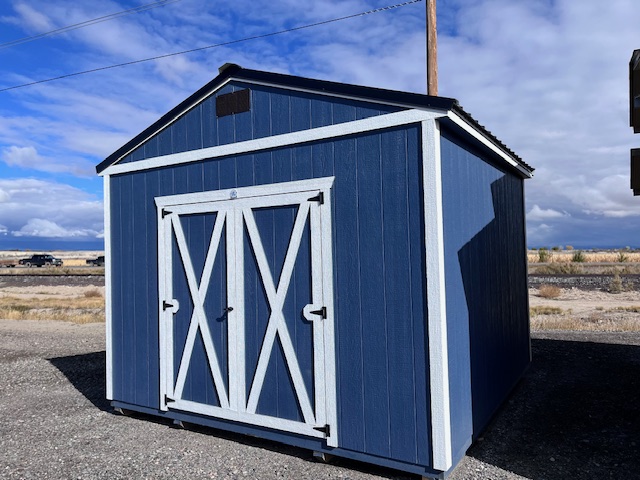 A blue and white shed with double doors stands on a gravel area against a partly cloudy sky. A wooden utility pole is visible behind it, and there is open land in the background.
