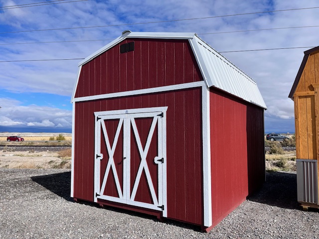 A red barn-style shed with white trim stands on a gravel lot under a partly cloudy blue sky. Power lines and a road are visible in the background along with grass and another wooden structure.