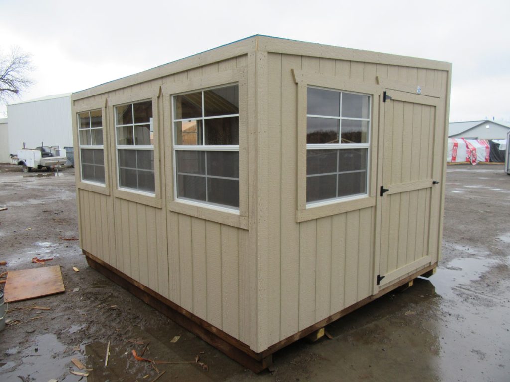 Beige wooden shed with four windows and a single door, positioned on a dirt ground. Nearby, some industrial buildings and equipment are visible in the background. Overcast sky above.