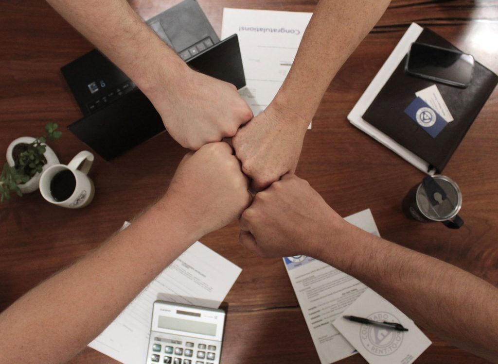 Four hands form a fist bump over a wooden table adorned with documents, a calculator, a phone, a mug, and a notebook. This scene symbolizes teamwork and collaboration. For more information or questions about our services, please contact us.