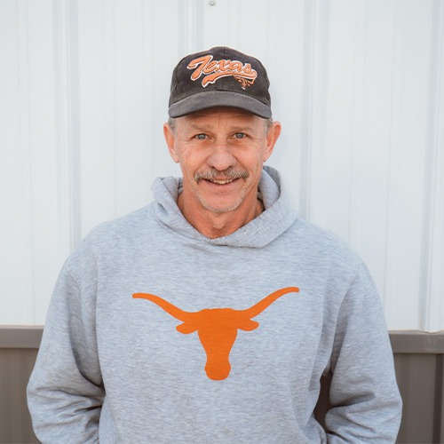 Jimmy with a mustache is wearing a gray hoodie with a burnt orange longhorn logo and a black cap that says "Texas" in orange. He is standing in front of a corrugated metal wall, smiling at the camera.