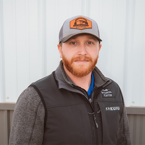 Matthew with a red beard wearing a gray baseball cap and a black KUIU vest over a gray jacket stands in front of a light-colored wall.