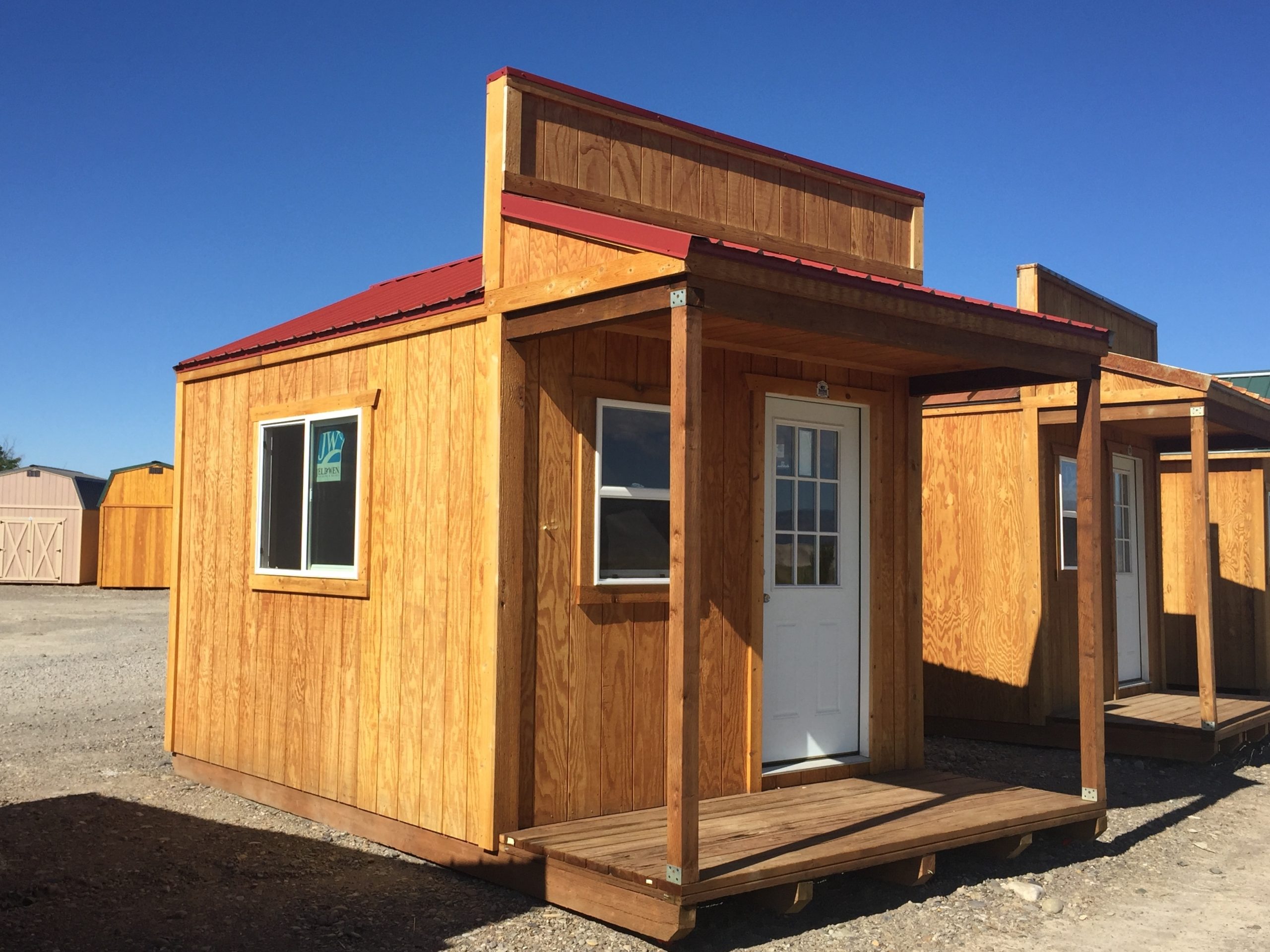 The country cabin boasts a charming small porch, featuring a red roof and a white door with a windowpane. This wooden haven sits gracefully on a gravel lot under the clear blue sky.
