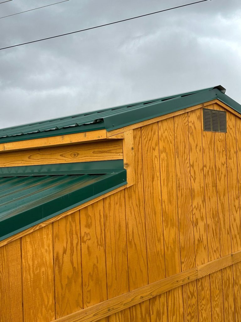 Side view of a yellow wooden building with a green metal roof under a cloudy sky. The wood features vertical planks, and there is a small vent near the roof line. Power lines are visible in the background.