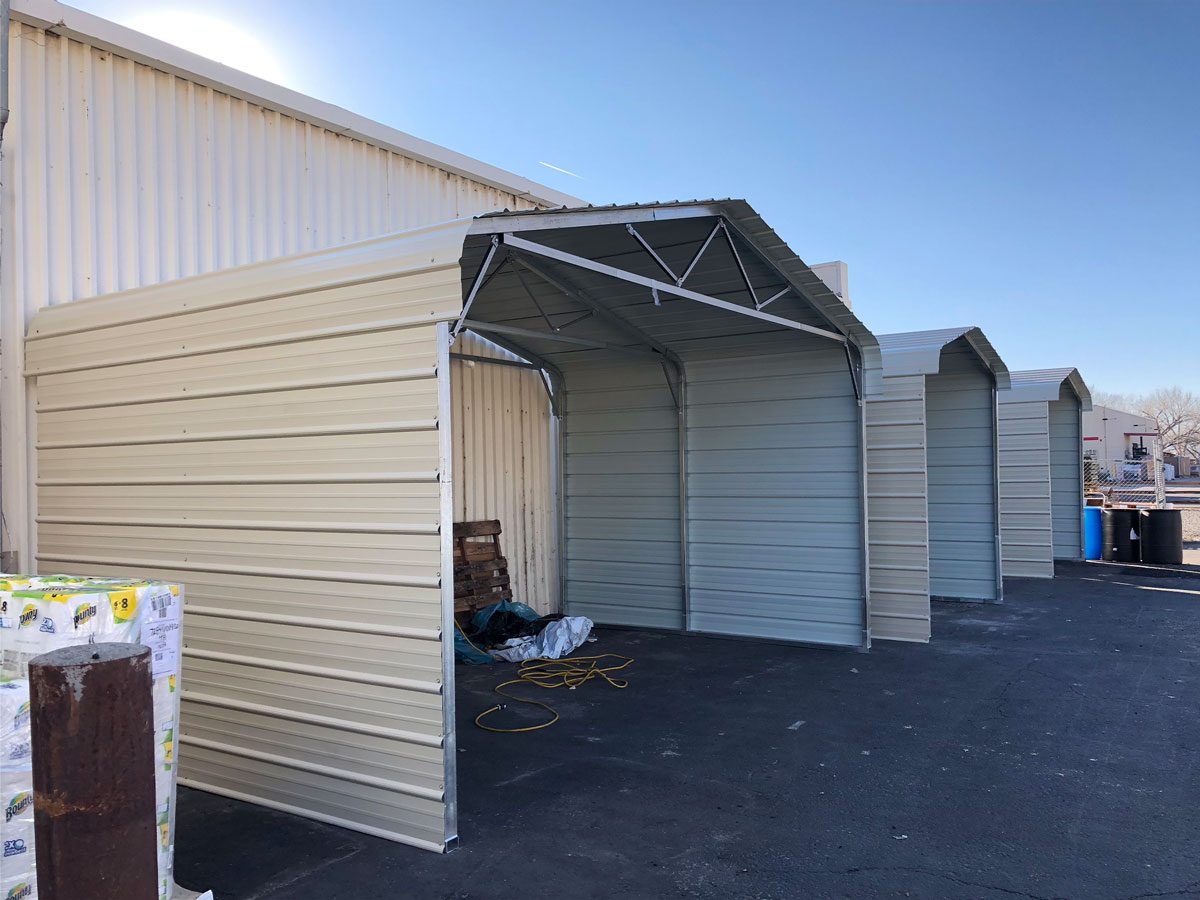 Three classic metal carports stand side by side on a concrete surface next to a beige industrial building. Each classic carport, with its peaked roof, is open at the front. A hose and some materials are visible nearby under one structure.
