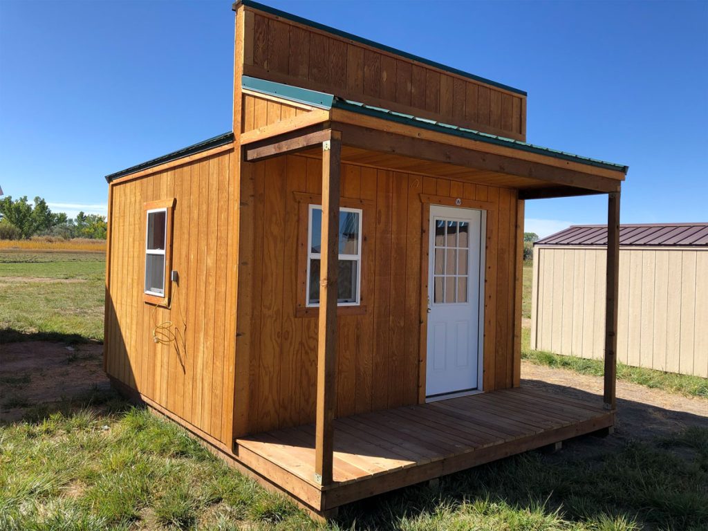 A small country cabin with a white door and front porch features a flat roof with a green edge. Set on lush grass alongside a similar structure, it stands under a clear blue sky, perfectly capturing rustic charm.
