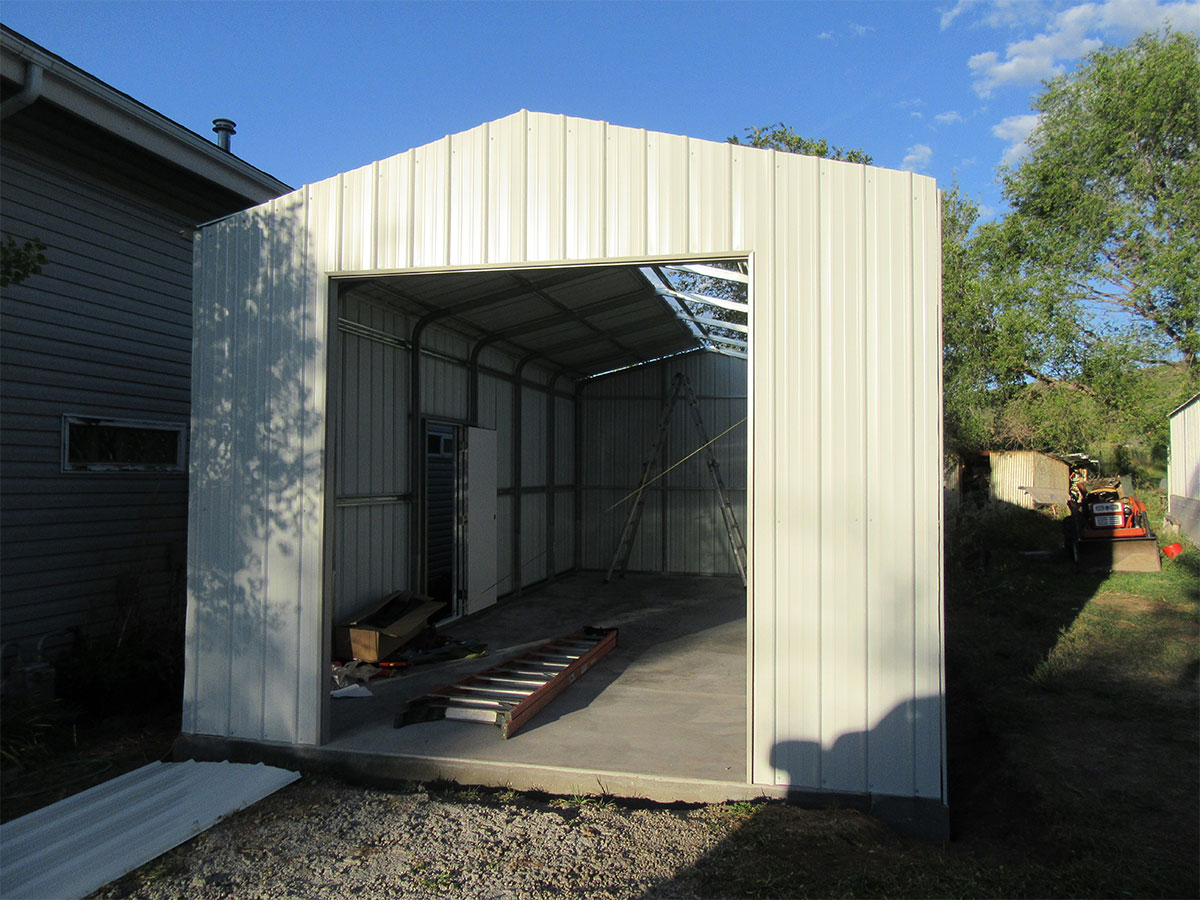 A partially constructed white metal frontier carport with an open front. Inside, there's a ladder, some scattered tools, and a concrete floor. Trees and a smaller building are visible in the background under a blue sky.