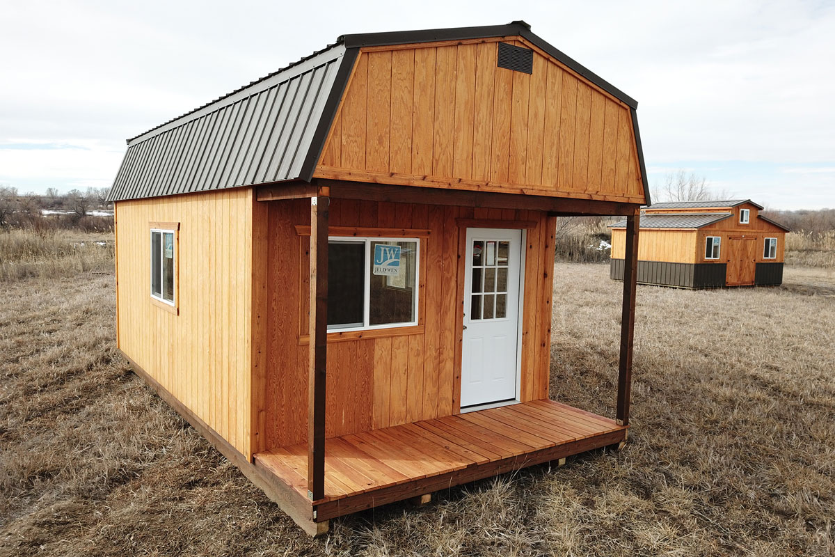 A small wooden cabin with a front porch and a white door sits on a grassy field. This lofted Dutch cabin features a metal roof and a single window on the side. Another similar cabin is visible in the background under a cloudy sky.