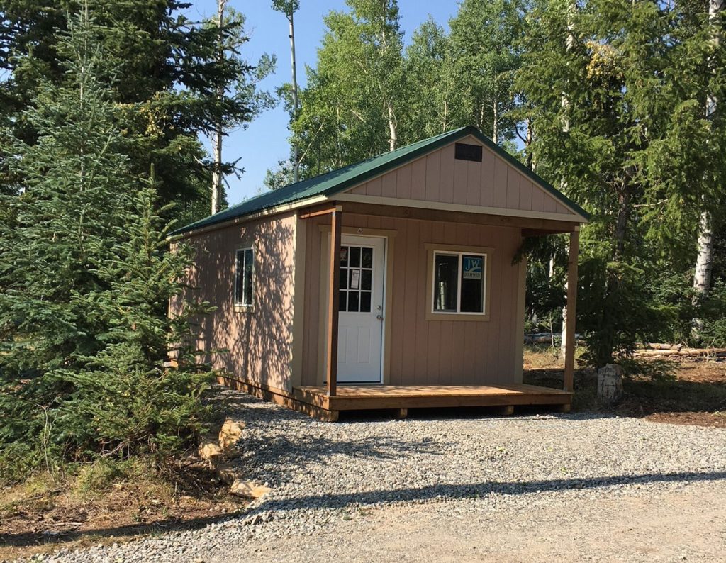 A small mountain cabin with a green roof and front porch is nestled among tall trees. It sits on a gravel path, surrounded by lush greenery under a clear sky. The door and window provide a cozy, inviting appearance.