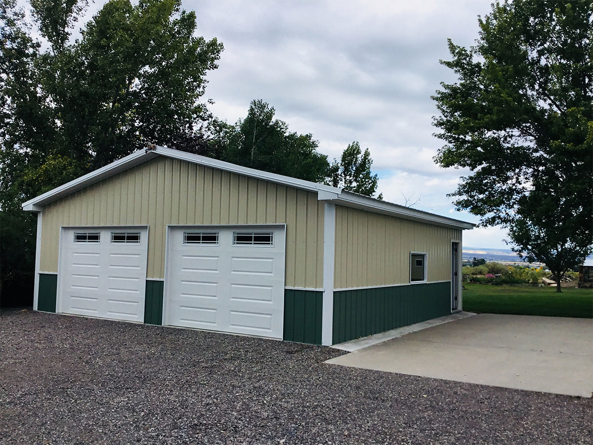 A beige and green metal summit carport, with two closed white doors, is surrounded by gravel and a concrete driveway. Trees and a grassy field stretch in the background under a cloudy sky.