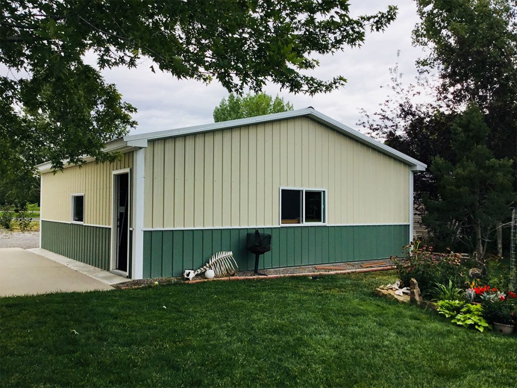 A small, light green and dark green metal summit carport is surrounded by lush greenery. It features a single window and side door. Nearby, a barbecue grill complements the neatly manicured lawn and flower garden under a partly cloudy sky.
