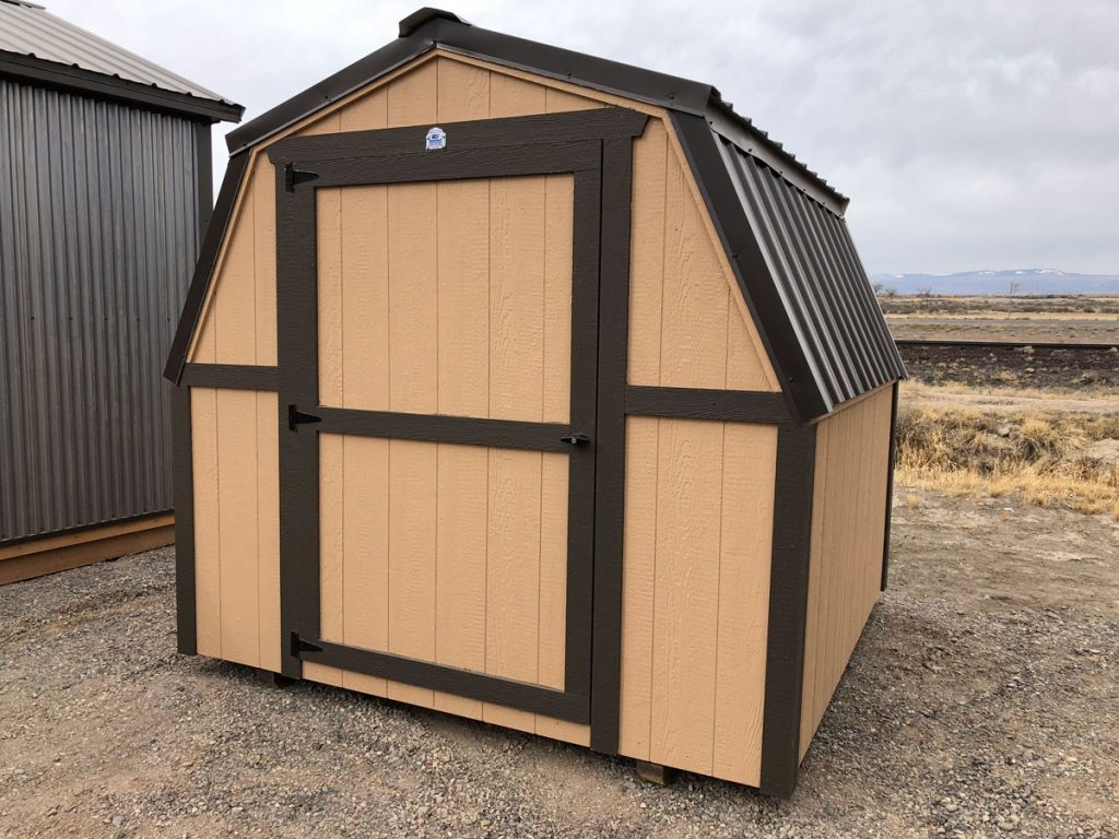 A small tan and dark brown mini barn with a single door, pitched roof, and vertical siding, situated on a gravel surface with a dry landscape and cloudy sky in the background.