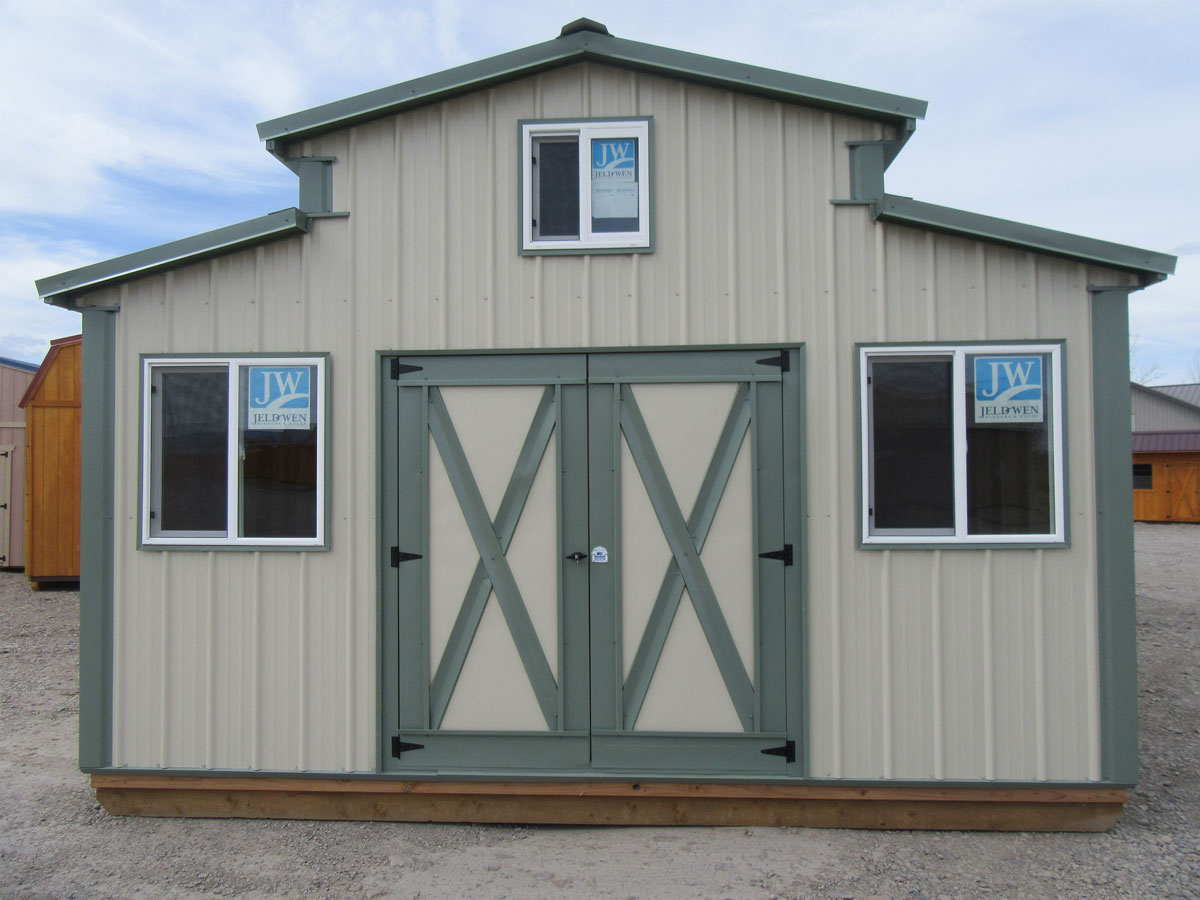 A light beige and green barn-style All American Shed with double doors and four windows, located on a gravel surface. The shed has a loft area with smaller windows, and "JW" logos on the upper windows.