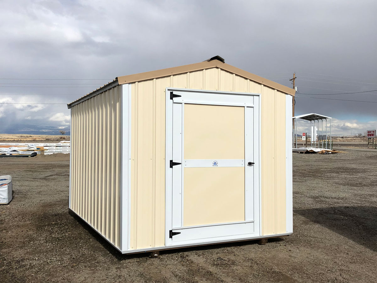A small beige and white utility shed with a single door, situated on a gravel lot under a cloudy sky. In the background, there are distant fields and a few scattered structures.
