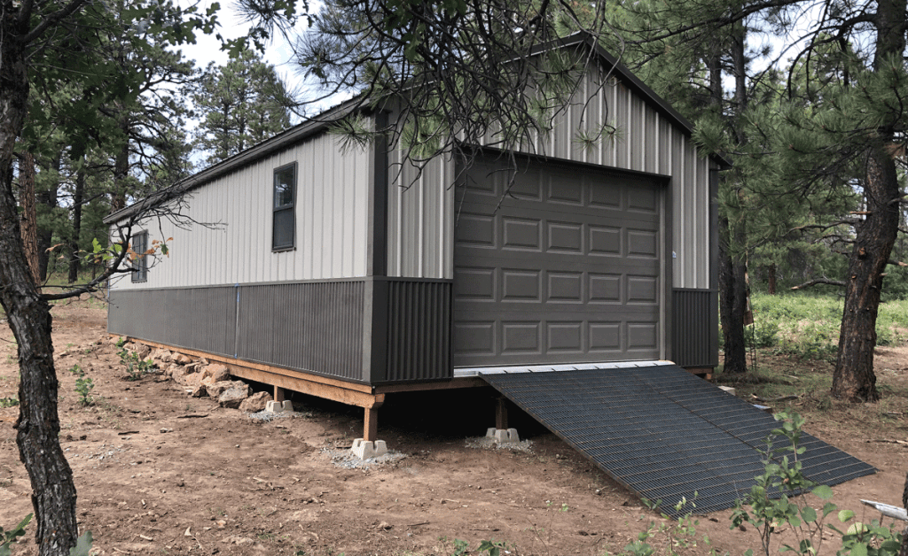 A gray, metal-clad Workshop with a brown garage door is set in a forested area. The structure is elevated on concrete blocks, and a ramp leads up to the door. Trees and grassy ground surround the shed.