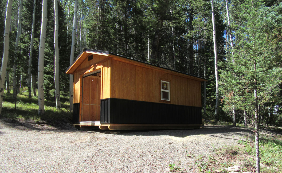 A small wooden Workshop with a black lower section sits on a gravel clearing, surrounded by tall trees and greenery in a forested area. A single window is visible on the side of the cabin, and the scene is illuminated by sunlight.