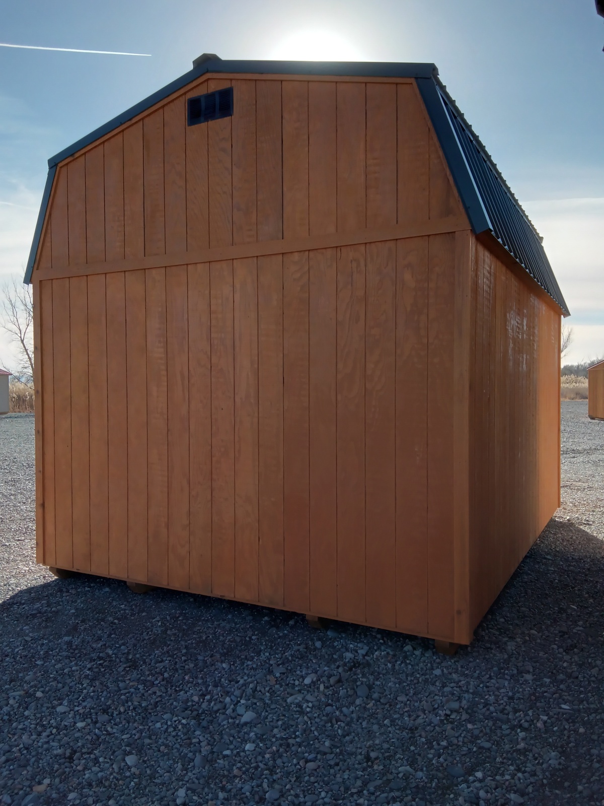 The Lofted Dutch Barn - 2383 features a classic barn-style roof and sits on gravel, with sunlight streaming from behind and soft shadows across its front. Above, the sky is mostly clear with a few wispy clouds.