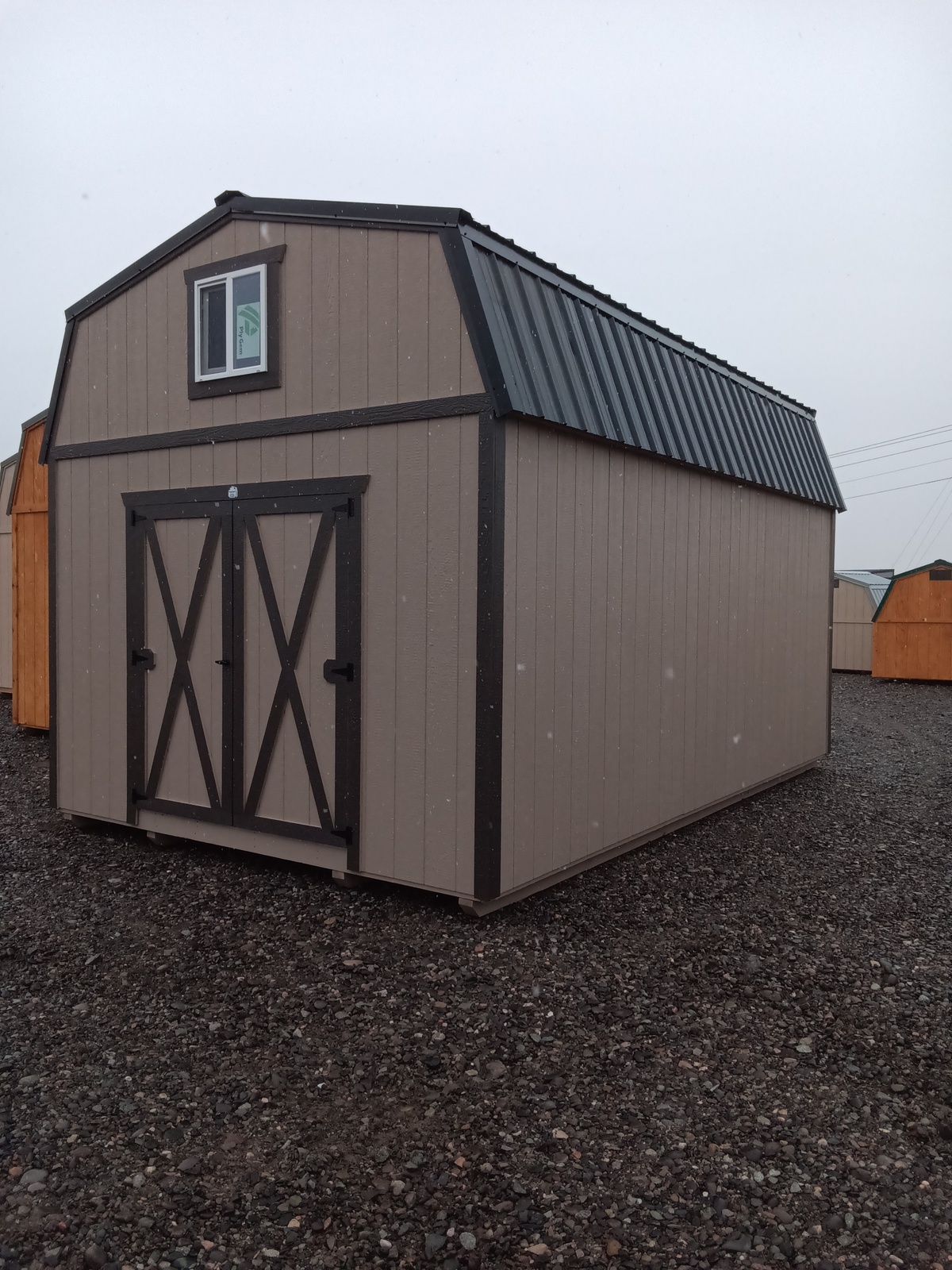 A Lofted Dutch Barn - 2382 sits on gravel, featuring a beige exterior with dark trim, a black metal roof, double "X" pattern doors, a small window above, and similar sheds visible in the background.