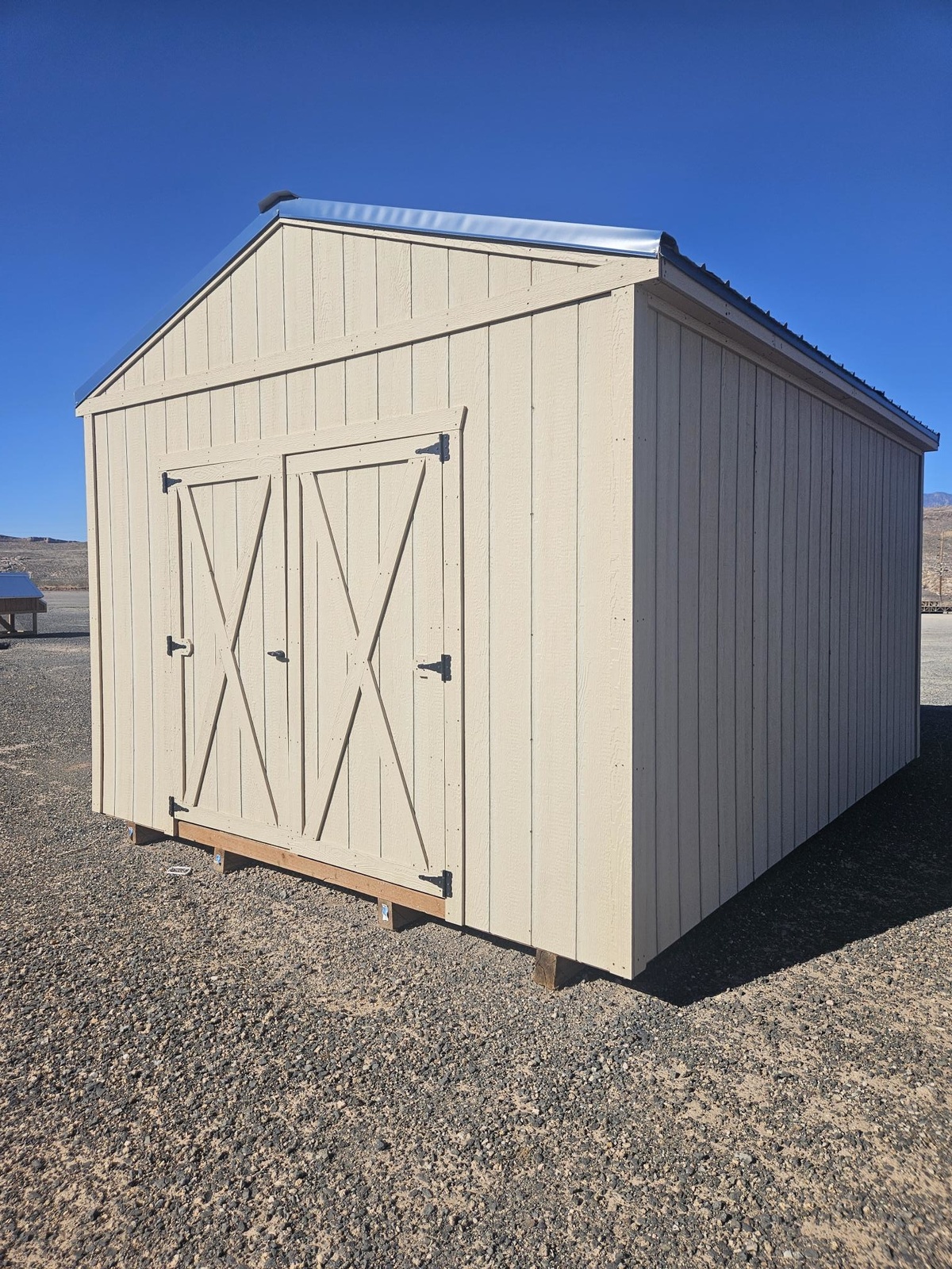The Classic Shed - 1718 is a beige wooden storage shed with double doors and a metal roof, situated on a gravel lot beneath a clear blue sky.