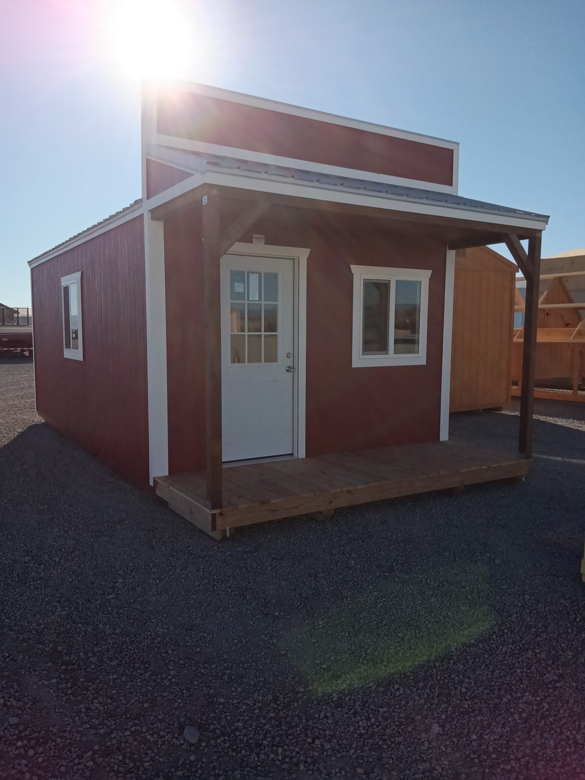 The Country Cabin - 2423 boasts a small red wooden exterior with a white door, two windows, and a covered porch, all set on a sunlit gravel lot.