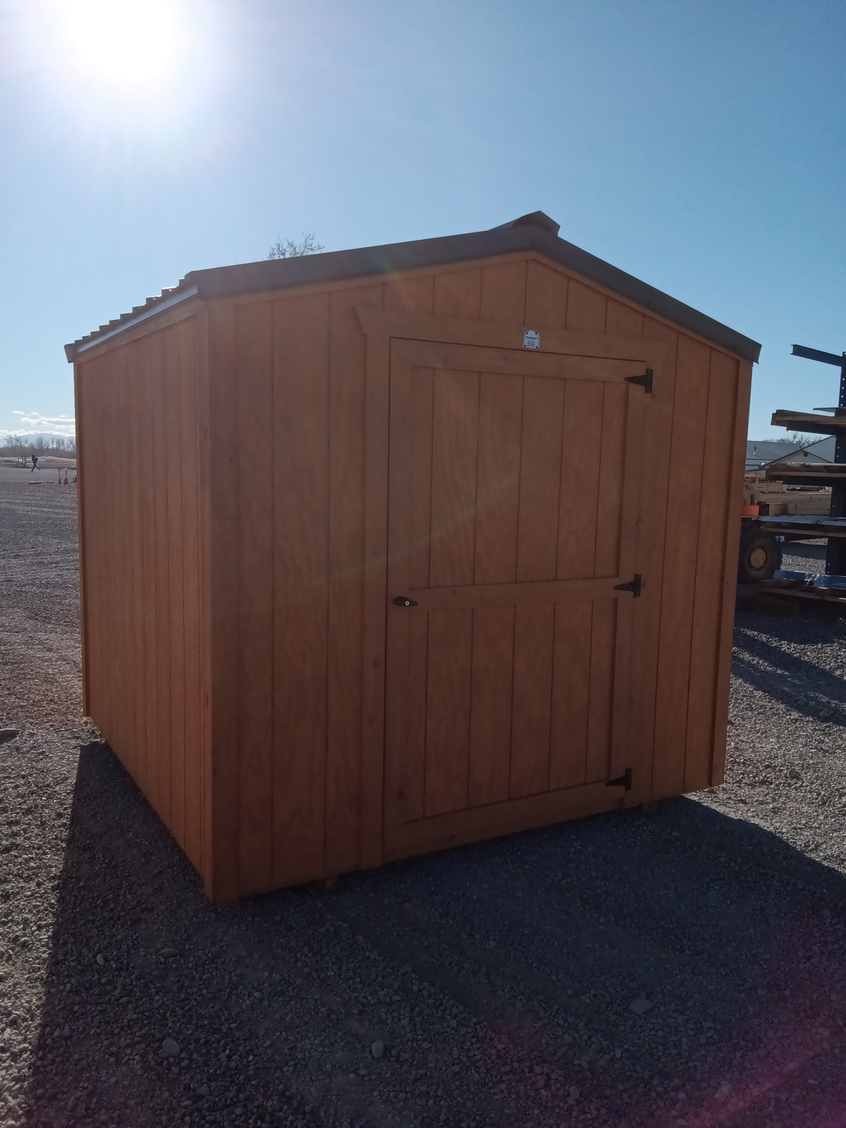 The Utility Shed - 2486, featuring a slanted roof and closed door, stands on a gravel lot under a clear blue sky with sunlight casting shadows across its front and side.