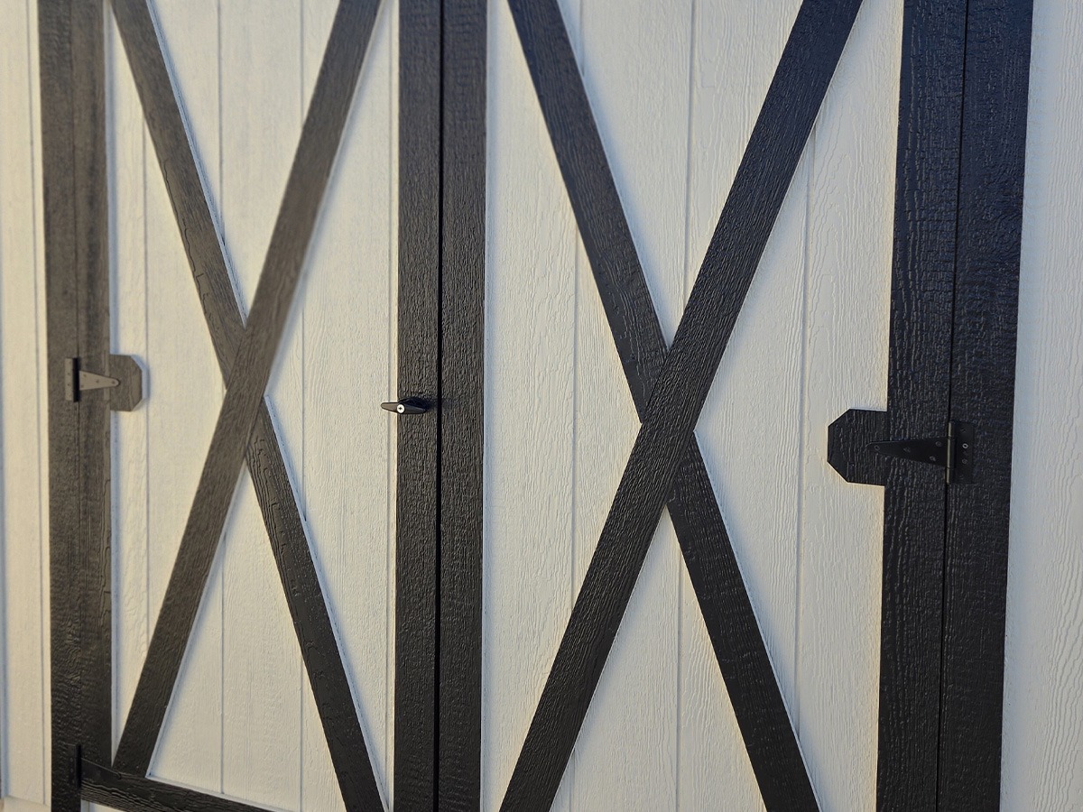 Close-up of a white wall resembling an All American shed, with diagonal and vertical black wooden beams arranged in a cross pattern. The beams share the appearance of door hinges or braces, casting shadows on the wall.