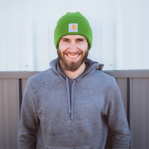 A smiling man with a beard, wearing a bright green beanie and a gray hoodie, stands among his team members in front of a white and gray wall.