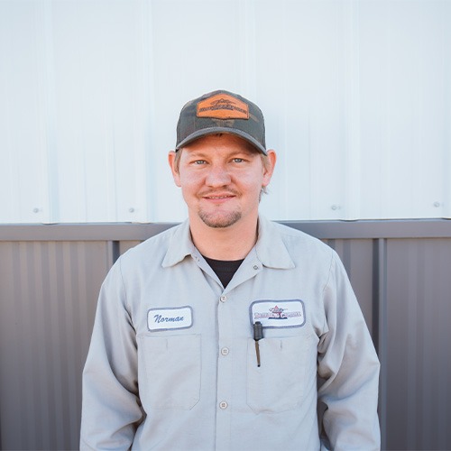 A man, one of the dedicated team members, stands in front of a metal building wearing a gray uniform and cap. His uniform features a name tag and logo. Smiling confidently at the camera, he embodies professionalism and teamwork.