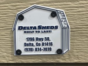 A sign shaped like a shed displaying the text: "DELTA SHEDS BUILT TO LAST! 1796 Hwy 50, Delta, CO 81416 (970) 874-7070," mounted on a textured surface.