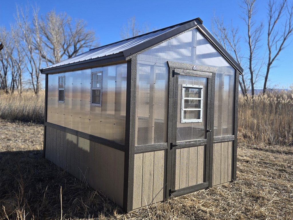 A small greenhouse with transparent walls and a dark frame stands like an architectural gem in a dry, grassy field under a clear blue sky. Leafless trees are visible in the background, juxtaposing natural beauty with the elegance of constructed buildings.