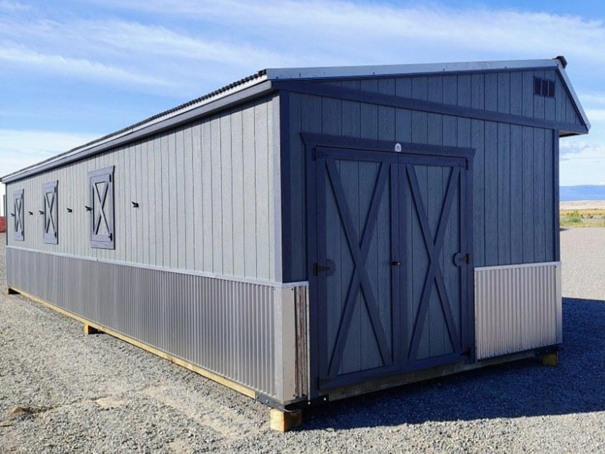 home-slide-1 A long, gray barn-like structure with double doors and small windows. The building has corrugated metal siding on the lower half and is situated on a gravel area under a clear blue sky.