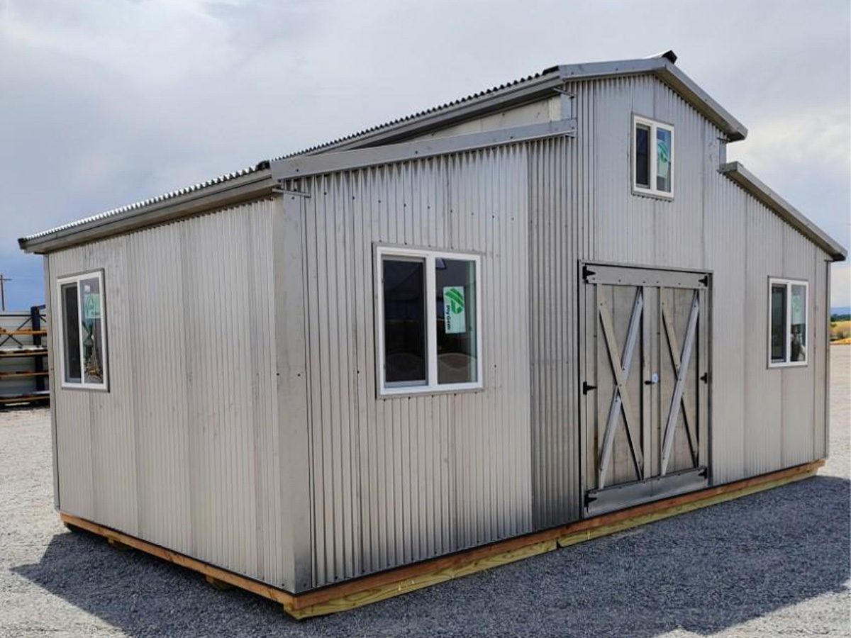 home-slide-2 A metal shed with a gabled roof and two windows on each side stands on a gravel lot. It has large double doors with a cross-brace design and corrugated metal siding. The sky is overcast in the background.