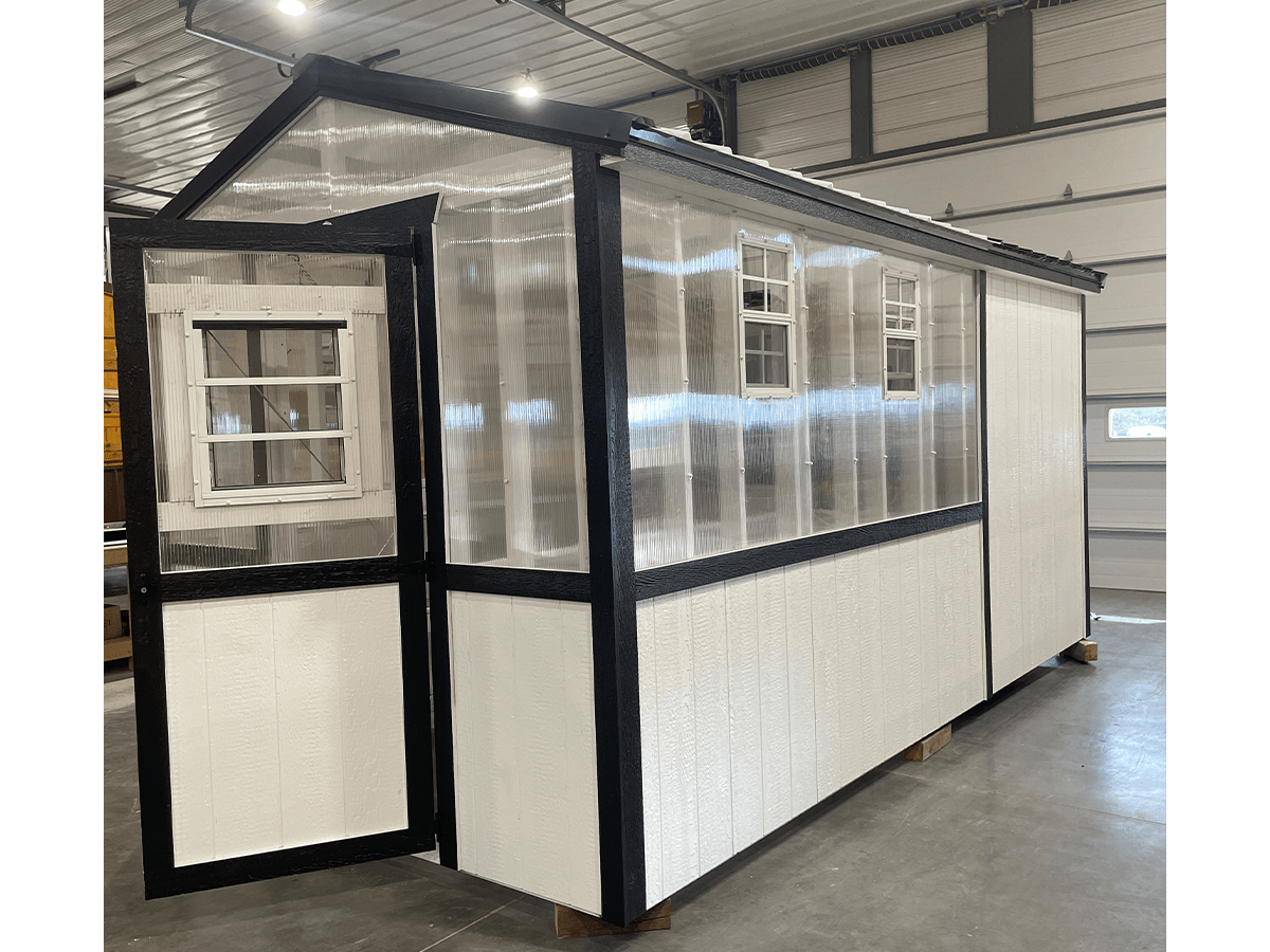 home-slide-3 Small, modular greenhouse with clear panels and black trim. It has multiple windows and a peaked roof, situated inside a large industrial space with concrete floors.