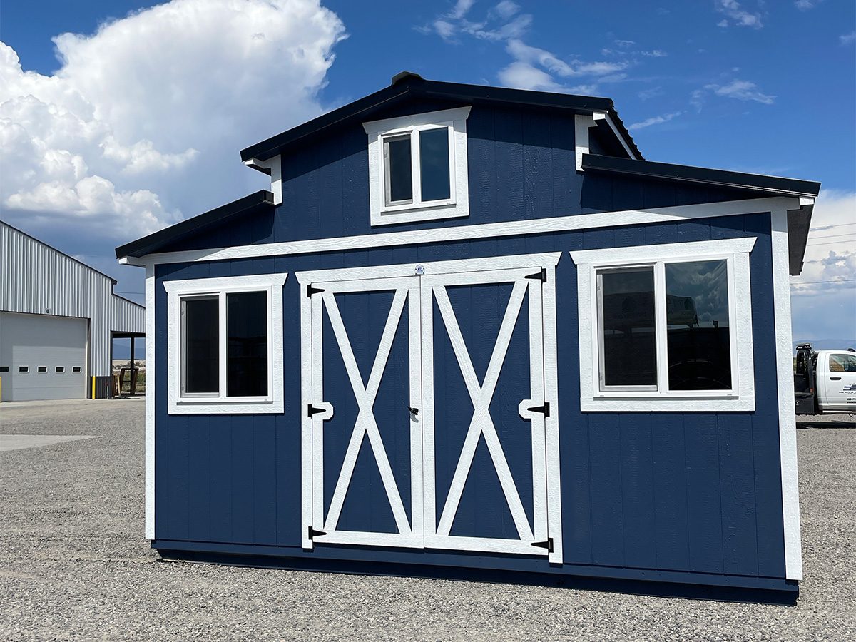home-slide-4 A blue and white barn-style storage shed stands on a gravel lot under a bright sky with fluffy clouds. It features double doors with a crossbuck design and multiple windows. A larger industrial building is visible in the background.
