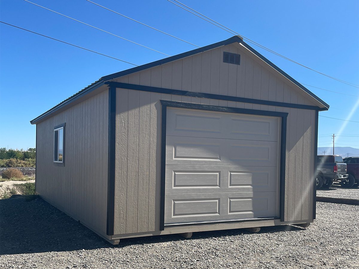 home-slide-5 A beige storage shed with a dark trim and a white garage door sits on a gravel surface under a clear blue sky. There's a small window on the side and some vehicles in the background.