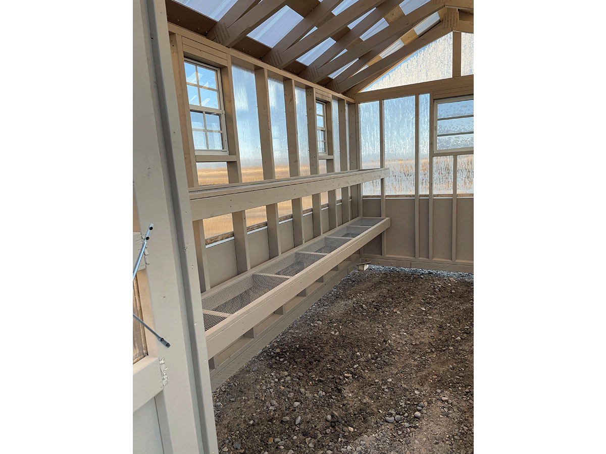 home-slide-6 Interior view of an empty greenhouse with soil flooring. It features wooden shelves and a transparent roof allowing natural light. The wooden structure has several windows for ventilation.