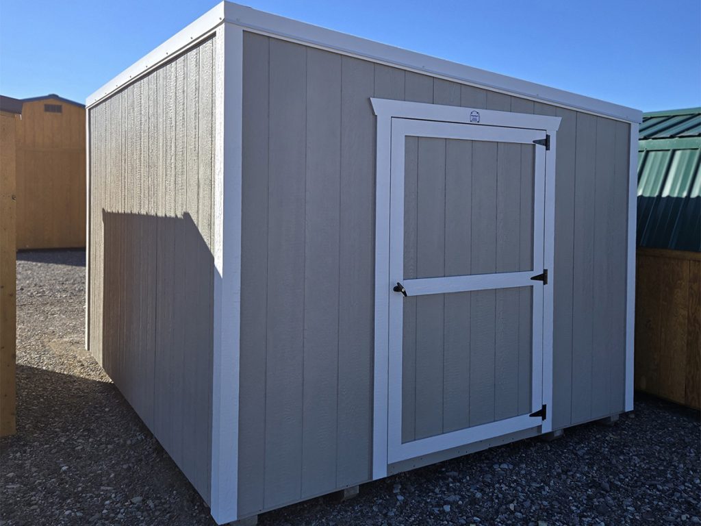 A gray lean-to shed with white trim and a single door stands on gravel under a clear blue sky, adding charm to the landscape of nearby buildings.