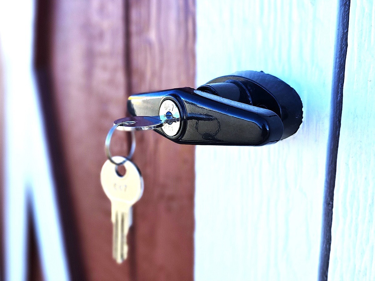A close-up reveals a shiny black door lock with a key inserted, the tag hinting at its All American Shed origin. The lock is mounted on a white door, framed by a wooden panel in the background, capturing an essence of classic craftsmanship.
