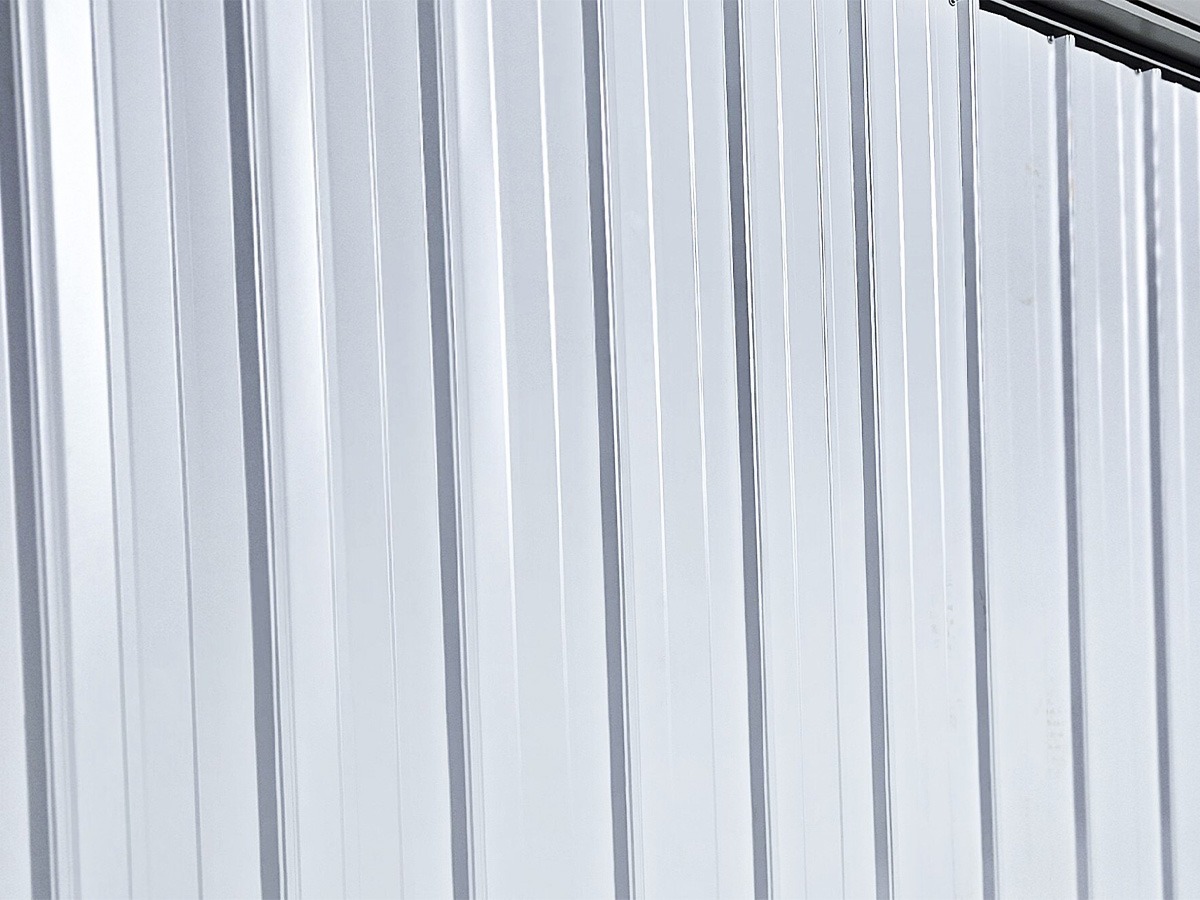 Close-up of an American shed's corrugated metal wall with vertical grooves, reflecting light. The metallic surface appears clean and evenly textured, with a slight gleam highlighting its smooth finish.