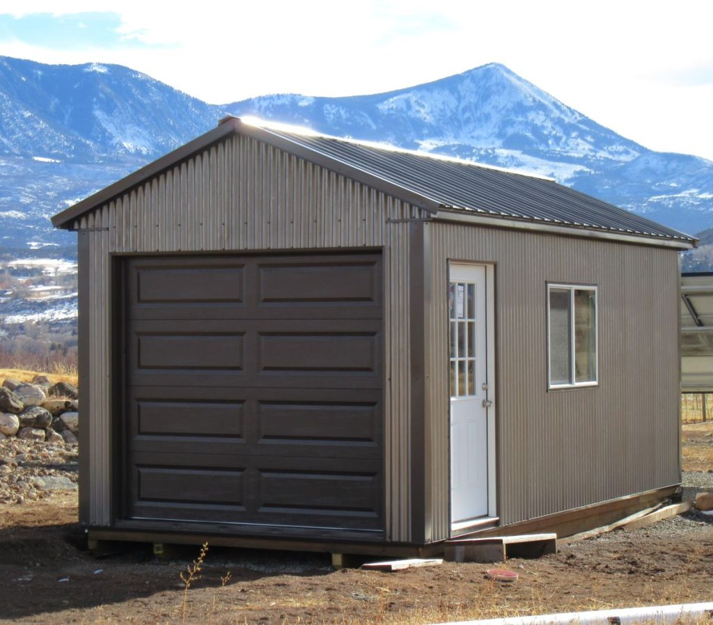 A small, brown metal shed with a white door and a roll-up garage door is set against a background of mountains under a clear sky. The structure has a window on its side and sits on a dirt ground.