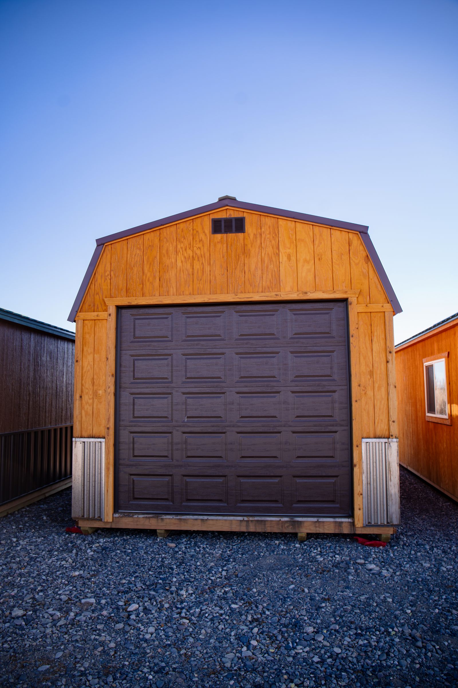 A small, brown wooden shed with a large garage door, set on a gravel surface. The shed is positioned between two similar structures under a clear blue sky.